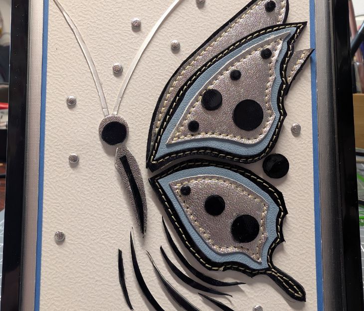 Tableau artisanal encadré avec un motif de papillon réalisé en cuir argenté, bleu clair et noir.
