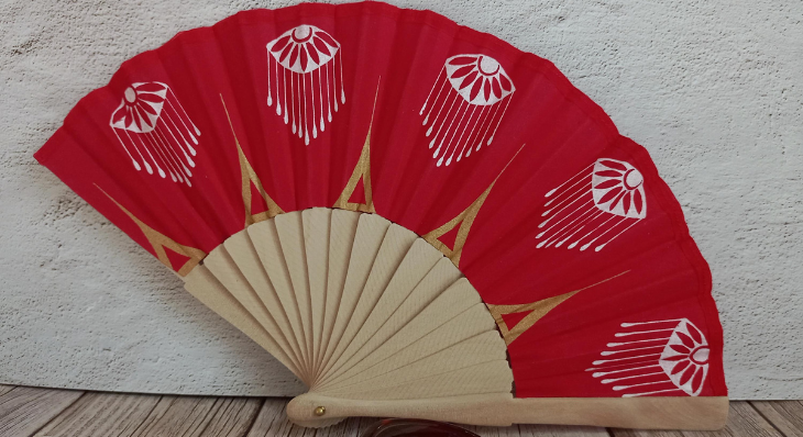 Éventail artisanal en coton rouge vif avec motifs sérigraphiés blancs et accents géométriques dorés.