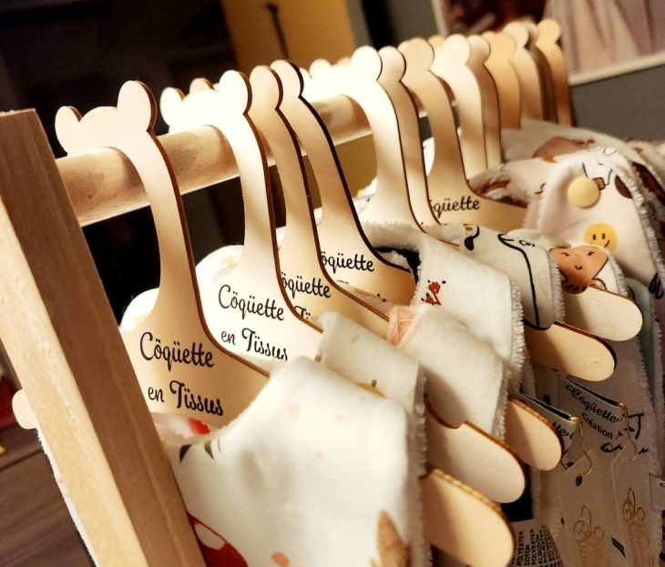 Collection de bavoirs artisanaux pour bébé présentés sur des cintres en bois gravés Côquette en Tissus.