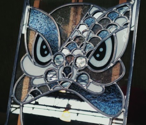 Détail d'un vitrail contemporain en plomb et verres bleus texturés représentant un masque féroce avec de grands yeux expressifs.